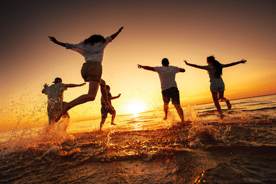 Silhouettes of big group of happy young friends are having fun, run and jump at calm sunset lake beach