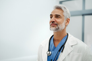 Portrait of confident mature doctor standing in Hospital corridor. Handsome doctor with gray hair wearing white coat, stethoscope around neck standing in modern private clinic, looking at camera.