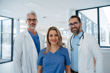 Portrait of confident three doctors standing in Hospital corridor. Medical team wearing white coat, stethoscope around neck standing in modern private clinic, looking at camera.