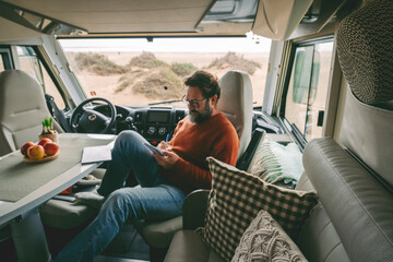 Handsome man with glasses sitting inside camper, writing and taking notes in notebook, smiling. Camping, outdoor activity, road trip, nature concept.