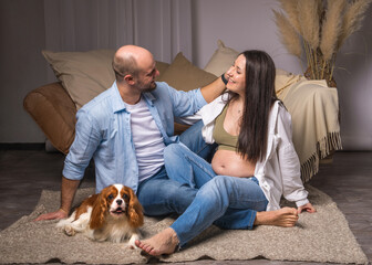 A man sits on the floor, a pregnant woman with a bare belly sits on the sofa. He holds small socks...