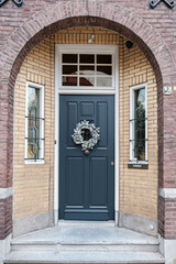 Elegance and classic design of dark, wooden front door adorned with a decorative wreath, set within arched entrance of a brick building. The door is complemented by two small windows on either side