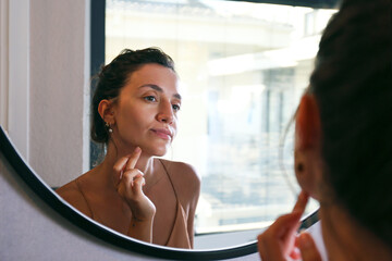 Young Middle Eastern woman wearing a silk nightdress, looking at the mirror and doing face...