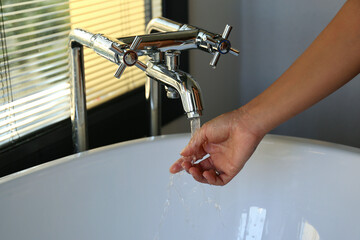 Cropped shot of young woman's hand touching the water flow from the double handle faucet to check the temperature. Close up, copy space, background.