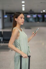 Asian woman with luggage using phone at a transportation arrival hall. 