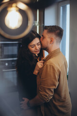 Married couple in the kitchen. A girl and a guy hugging. The man holds the girl by the waist and kisses her temple. Blurred kitchen background with stove and highlights in the foreground.