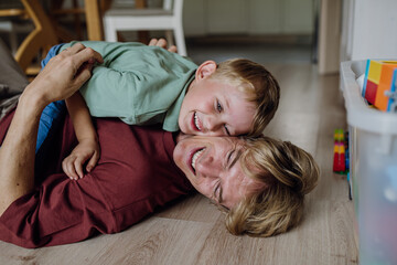 Portrait of father embracing his son at home, having fun, lying on floor and laughing.