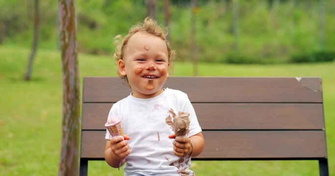 Cheerful and expressive little one sits on bench in park, laughing and enjoying melted ice cream from two cones. His face and stained t-shirt reflect genuine joy and fun of this playful moment