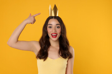 Beautiful young woman pointing at princess crown on yellow background