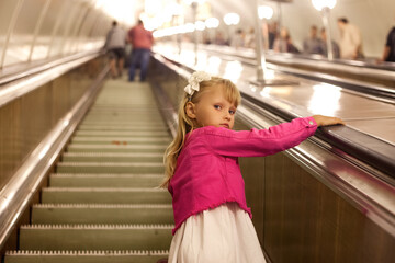 Lovely child girl 5 year old going up subway escalator in underground metro, looking around at camera. Kid passenger in pink jacket at subway. Public urban transportation concept. Copy ad text space