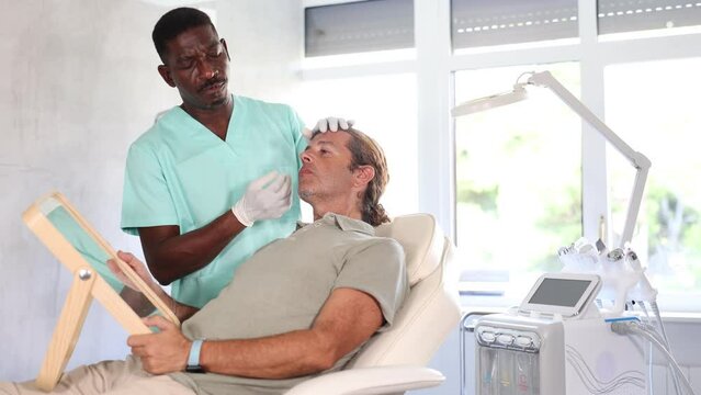 Interested middle-aged patient of medical aesthetic clinic sitting on examination couch and looking in mirror while professional African American cosmetologist 