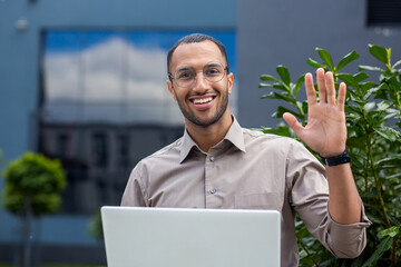 Smiling African American professional outdoors with laptop, greeting someone near an office campus.