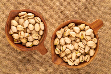 A small amount of roasted pistachios in two wooden cups on a jute cloth, macro, top view.