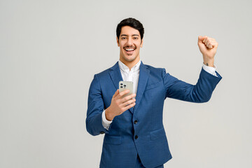Happy businessman in sharp blue suit celebrates success with a fist pump while holding a smartphone in his other hand, against a neutral gray background, positive news communicated via mobile.