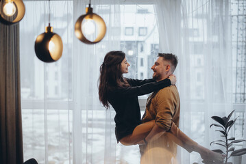 Young couple in a hotel near the window. The guy holds the girl in his arms. The girl holds her hands on the guy's neck. They smile. The city is blurry in the background. Glare in the foreground
