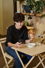 Fototapeta na wymiar happy and curly young man holding plant-based tofu burger texting on smartphone in vegan cafe