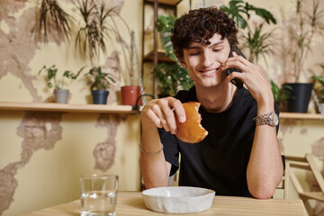 Fototapeta na wymiar happy and curly young man holding plant-based tofu burger and talking on smartphone in vegan cafe