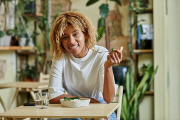 happy african american woman in braces eating fresh salad in bowl inside of plant-filled vegan cafe