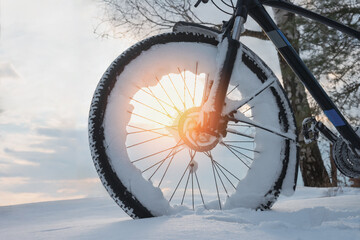 Close-up of a snow-covered wheel of a standing bicycle in the light of the setting sun in the...