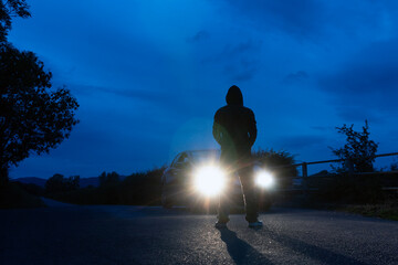A mysterious hooded figure, silhouetted against the headlights of a parked car. On a lonely spooky country road at night.