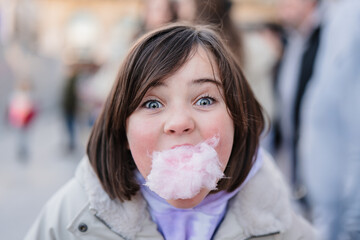 Close-up of excited girl with wide eyes looking at camera while enjoying a mouthful of cotton candy on a city blurred street background