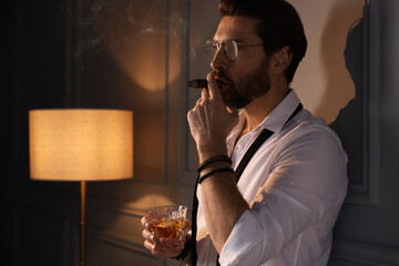 Handsome man with glass of whiskey smoking cigar at home. Space for text