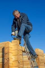 a man drills a hole in a beam with a drill, builds a wooden house made of profiled timber against a blue sky background - 715567586