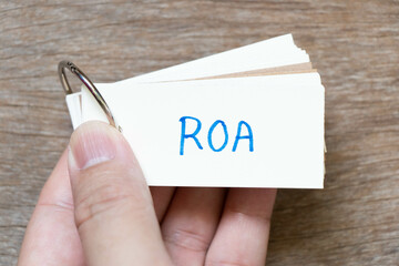 Hand hold flash card with handwriting in word ROA (Abbreviation of Return on assets) on wood...