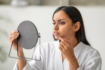 Unhappy eastern young woman in white robe looking at mirror