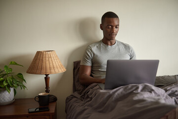 Young African man using a laptop in bed in the morning