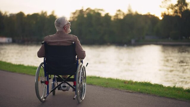 Man in wheelchair walking in city park. Senior male breathing fresh air near pond admiring enjoying a nature at sunset. Live with physical disability, special needs people in old age concept.