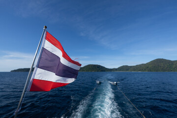 A Thai flag blows in the wind while sailing on a ship. Two dinghies are towed, an island in the...