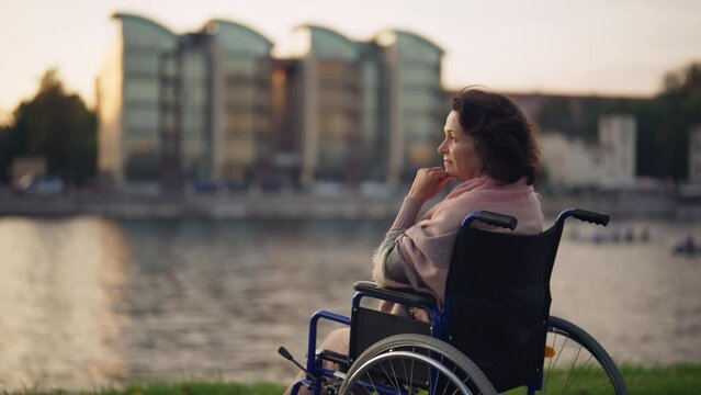 Woman in wheelchair walking in city. Senior mature pensive woman breathing air outdoor thinking looking around enjoying pastime in evening. Live with physical disability, special needs people concept.