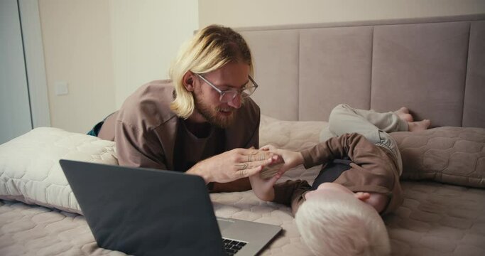 A happy blond man in glasses with a beard plays with his little albino son with white hair color in rock paper scissors and then turns on a cartoon on a gray laptop while spending time together in the