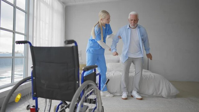 Nurse helps old wheelchair man to walk. Physical therapist woman provides medical help support to senior male training studying to go. Recuperation after trauma, temporary paralysis concept.