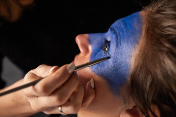 Close-up portrait of woman having her face painted blue by an artist with brush. Creative makeup, performance makeup - obrazy, fototapety, plakaty