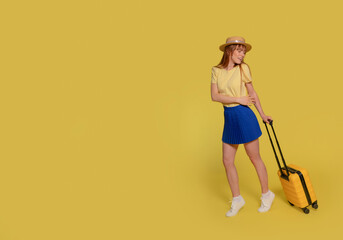 a happy young woman in blue skirt, a yellow T-shirt, and hat carrying a suitcase on a yellow...