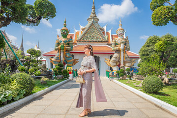 Young woman wearing traditional Thai dress stands holding an antique bag with giant front ornaments at the Wat Arun Temple, a popular destination for tourists around the world. Bangkok Thailand