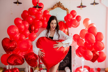 Young woman smiling while standing in room decorated for celebration Valentines Day. Girl holds large red heart shaped balloon. Rose petals scattered on floor along with pillows gift boxes. Mock up