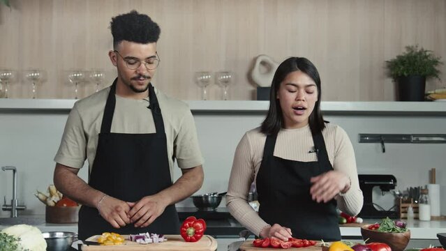 Whip zoom shot of young male and female chefs in aprons standing in kitchen, demonstrating vegetables and telling about food ingredients for recipe while hosting cooking show