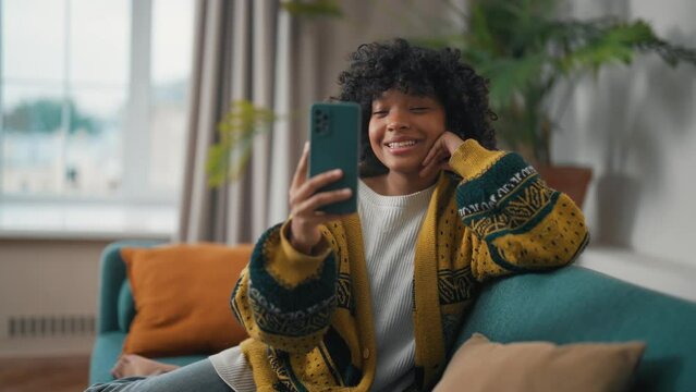 Young woman with phone talking with friends. African American girl hold in hand use mobile phone. Teenager relax spend free spare time in living room with phone. Communication on social networks
