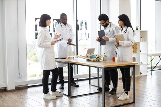 African man medical teacher teaching anatomy to university students or young doctors. Arabian man, Caucasian and Indian women standing near table, working with human skeleton model with body organs. - Powered by Adobe