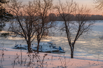 A small ship and boats on a small pier, covered with snow, on a river covered with ice and snow.