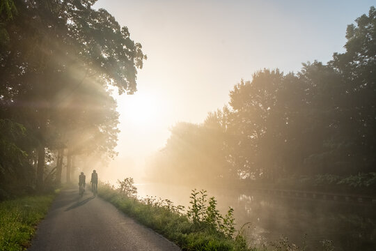This image captures a tranquil morning scene with two individuals walking along a riverside path. The rising sun, barely seen through the mist, creates a soft, golden glow that envelops the scene. The