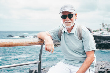 Portrait of senior traveler man with cap and sunglasses sitting at sea, elderly white haired...