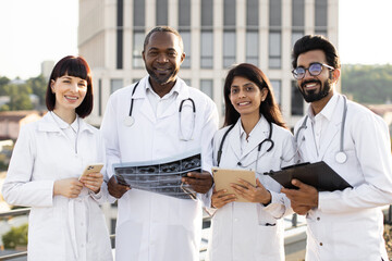 Close up of successful team of multinational doctors in white coats with stethoscopes holding gadgets with results of a patients examination in hands, looking at camera. - obrazy, fototapety, plakaty