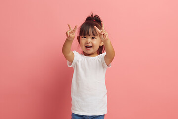 little asian etnicity funny girl showing V gesture with two hands standing over pink isolated...