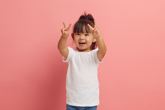 little asian etnicity funny girl showing V gesture with two hands standing over pink isolated background. 