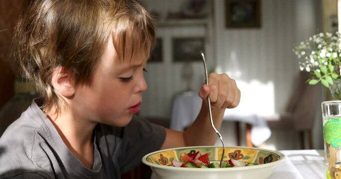 Portrait of a boy seven years old eating vegetable salad while sitting at home in cozy sunlight healthy habit side view
