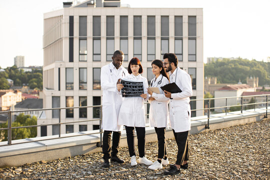 Team of four professional male and female multiracial doctors examining patient's tomography scans while standing outside the modern hospital building. Doctors discussing x-ray image.